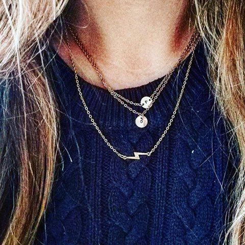 Gelaagde gouden ketting set met twee initiaal bedels en een bliksemflits hanger gedragen op een donkerblauwe kabeltrui, beschermende amulets van lux & luz.
